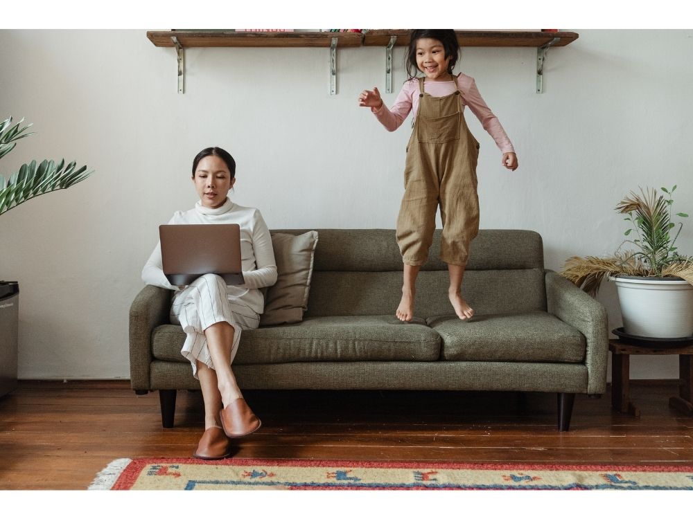 mulher pesquisando na internet com uma menina pulando no sofá 