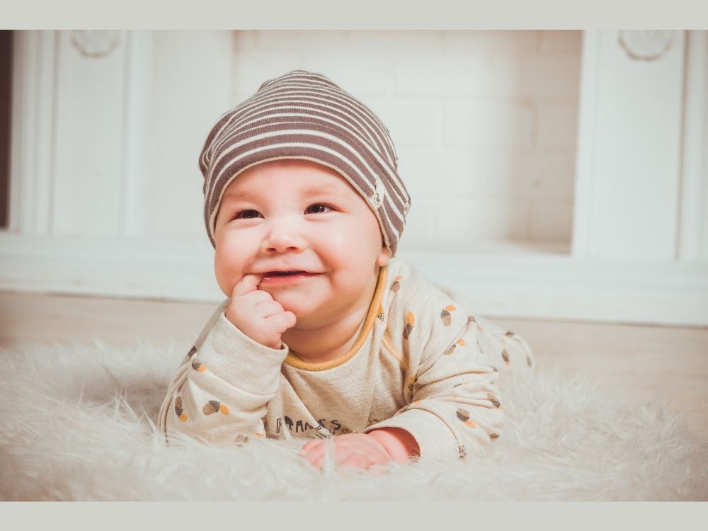 bebê branco com toquinha listrada sorrindo com dedo na boca