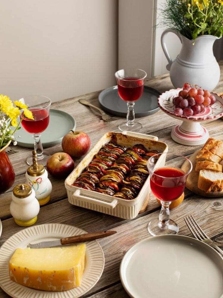 mesa posta com travessas de porcelana e taças com bebida dentro com maças e uvas em volta